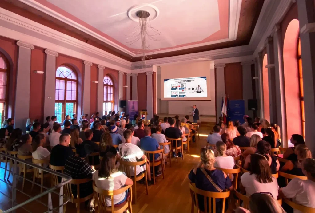 Audience seated in a large historic hall watching a presenter with a slide projected on a screen at the front of the room.